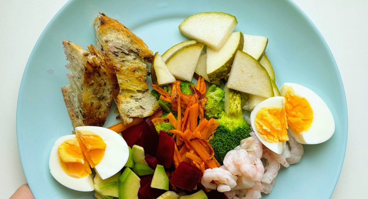 Assiette bleue avec une composition équilibrée d'œufs durs, crevettes, brocoli, carottes, avocat, betterave, poire et pain, illustrant les repères d'une alimentation saine