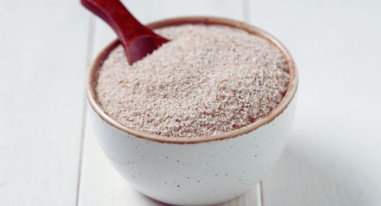 Bol blanc rempli de poudre d'enveloppe de psyllium avec une cuillère rouge, illustrant ce complément alimentaire riche en fibres aux nombreux bienfaits digestifs